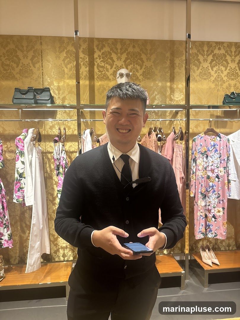 A smiling male sales associate stands in a brightly lit boutique, holding a smartphone. He is wearing a black cardigan over a white collared shirt and a black tie. Behind him, racks of clothing with floral patterns and solid colors are displayed, along with handbags and shoes on shelves. The walls are decorated with a gold damask pattern.