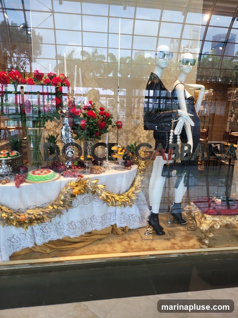 A store window display features two mannequins dressed in black, one wearing a leather jacket and the other a dress. They are accessorized with handbags and jewelry. In front of the mannequins, a table is laden with artificial fruit, a cake, and red roses. A gold garland drapes across the table. The store's name, 'DOLCE & GABBANA', is visible in large letters behind the display. The reflection of the sky and buildings is visible in the glass.