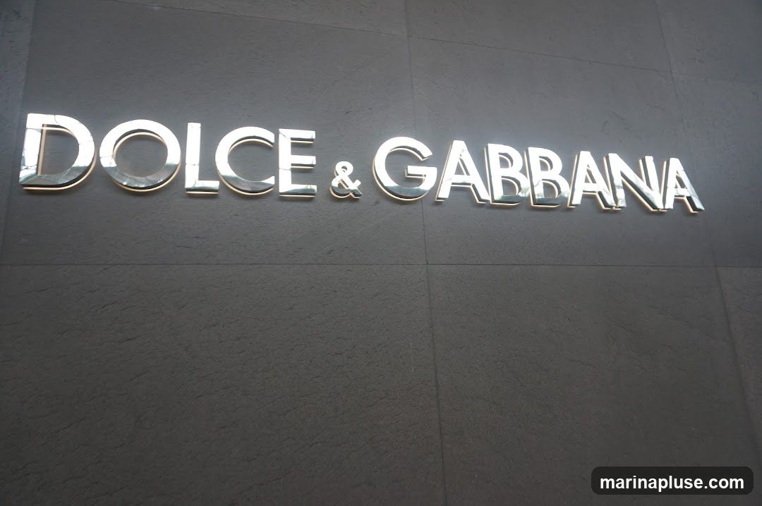 A close-up shot of the Dolce & Gabbana logo, rendered in large, metallic, three-dimensional letters against a dark, textured wall. The letters are polished and reflect light, giving them a sleek and luxurious appearance. The sign is mounted directly onto the building's facade.