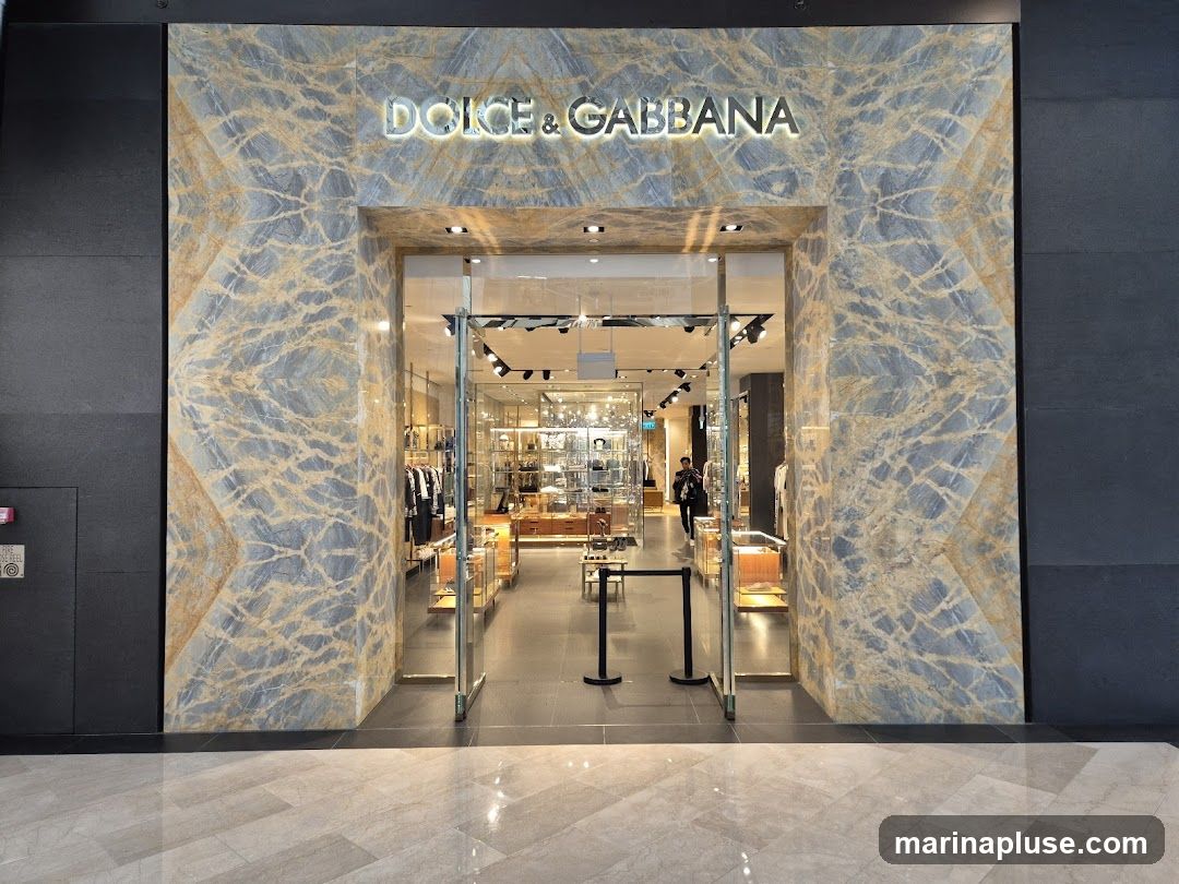 The exterior of a Dolce & Gabbana store. The entrance is framed by marble-patterned walls and glass doors. The store's name is illuminated above the entrance. Inside, clothing and accessories are displayed on shelves and racks.