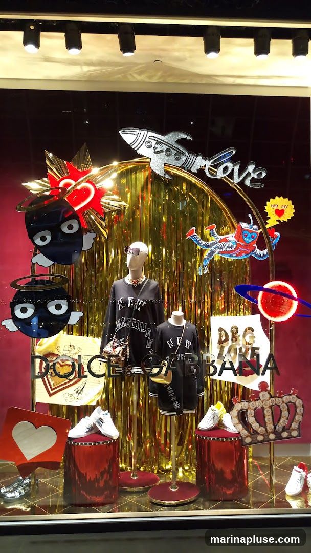 A festive and whimsical window display for Dolce & Gabbana, featuring mannequins dressed in black apparel with prominent branding. The display is adorned with space-themed elements like rockets and planets, alongside hearts and crowns, all set against a backdrop of shimmering gold fringe.