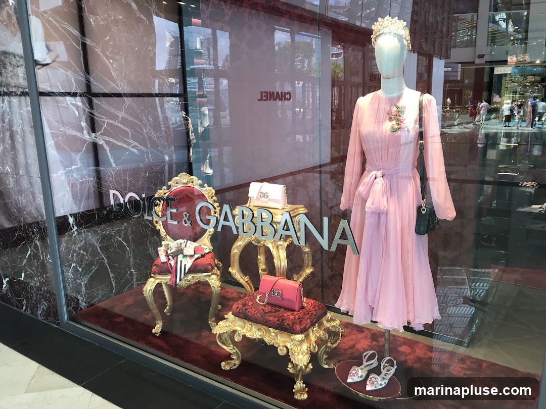 A storefront window display for Dolce & Gabbana. The display features a mannequin wearing a pink dress, a tiara, and a black handbag. To the left, there are ornate chairs and small tables displaying handbags and shoes. The store name "DOLCE & GABBANA" is visible on the window.