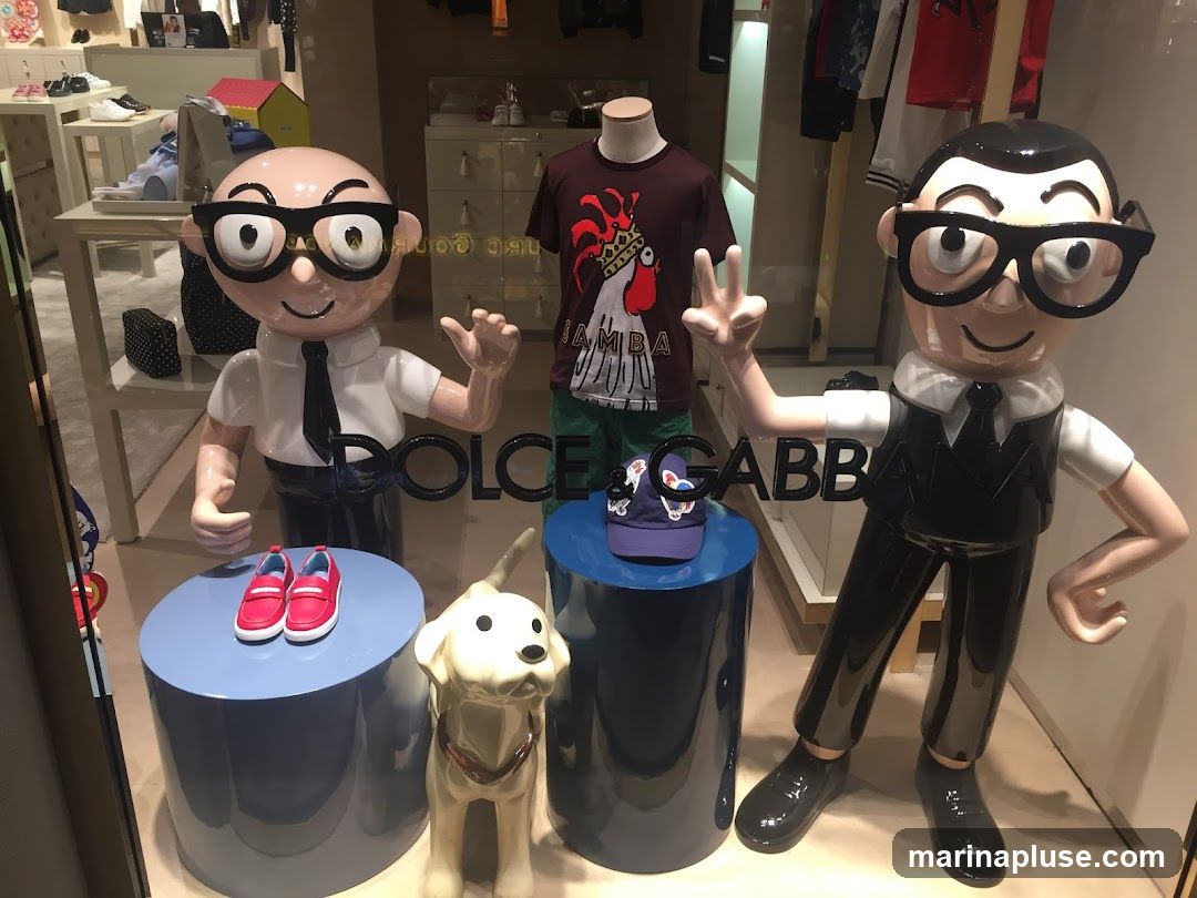A whimsical window display features two cartoonish mannequins with large glasses and ties, flanking a white dog figurine. The mannequins are dressed in formal attire. The left mannequin presents a pair of red loafers on a blue pedestal, while the right mannequin strikes a peace sign. Behind them, a t-shirt with a rooster graphic is displayed on a mannequin. The store's name, "DOLCE & GABBANA," is prominently visible in large black lettering across the display. The background shows clothing racks and shelves stocked with merchandise, suggesting a high-end fashion boutique.