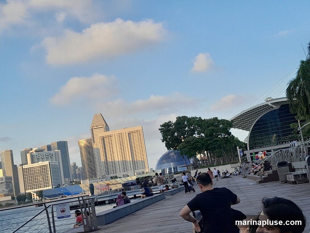 A wide shot of a waterfront promenade with people sitting and walking. In the background, a modern cityscape with tall buildings and a distinctive domed structure is visible. The sky is blue with scattered clouds. A large, arched building with the text "THE SHOPPES AT MARINA BAY" is prominent on the right side.