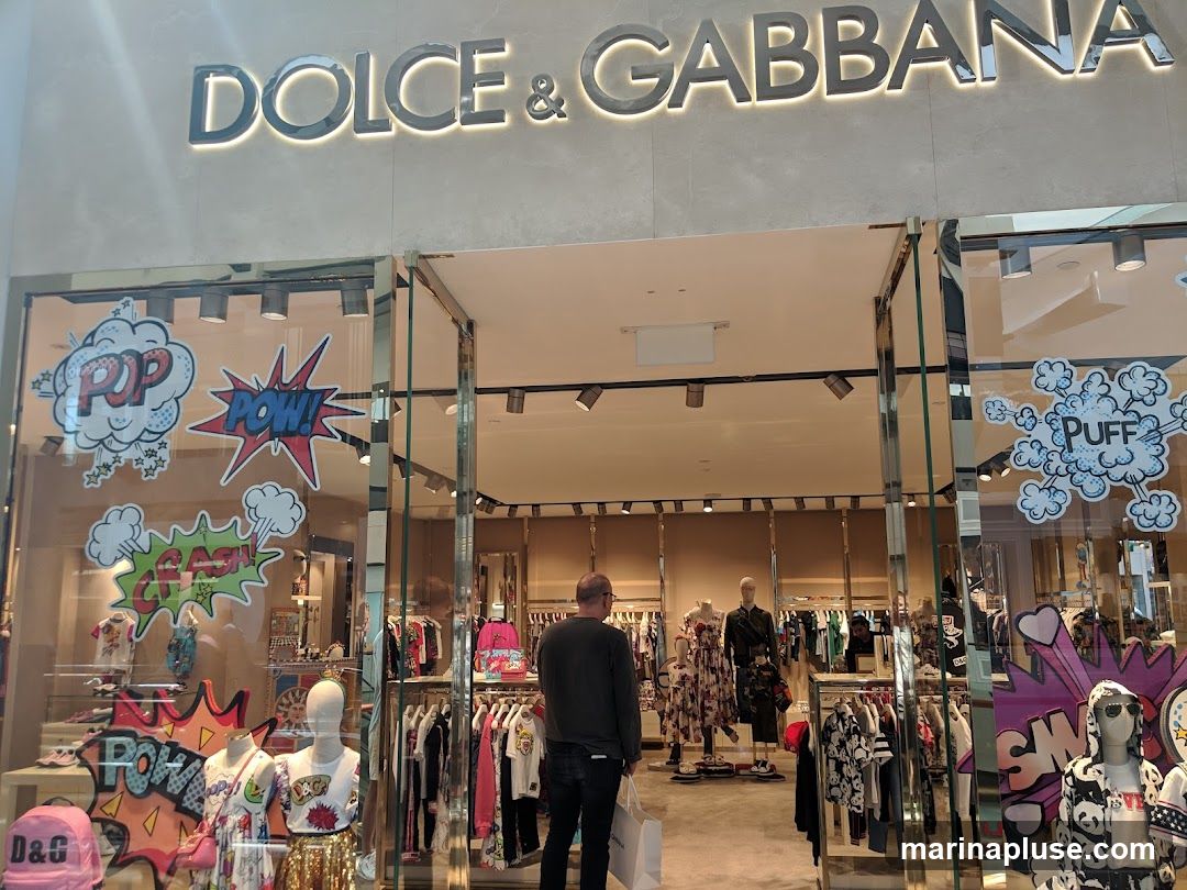 The exterior of a Dolce & Gabbana store. The large, illuminated "DOLCE & GABBANA" logo is visible above the entrance. The storefront windows display clothing on mannequins and racks, with comic-book-style graphics like "POP," "POW," "CRASH," and "PUFF" decorating the glass. A person is entering the store carrying a white shopping bag.