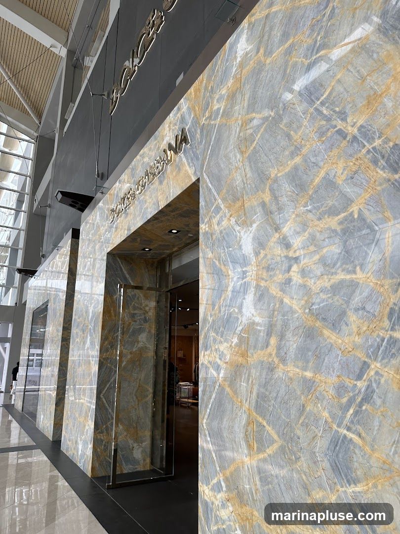 The exterior of a Dolce & Gabbana store, featuring a grand entrance with marble-patterned walls and illuminated signage. The store's name is visible in large, metallic letters above the entrance. Through the glass doors, racks of clothing can be seen inside.