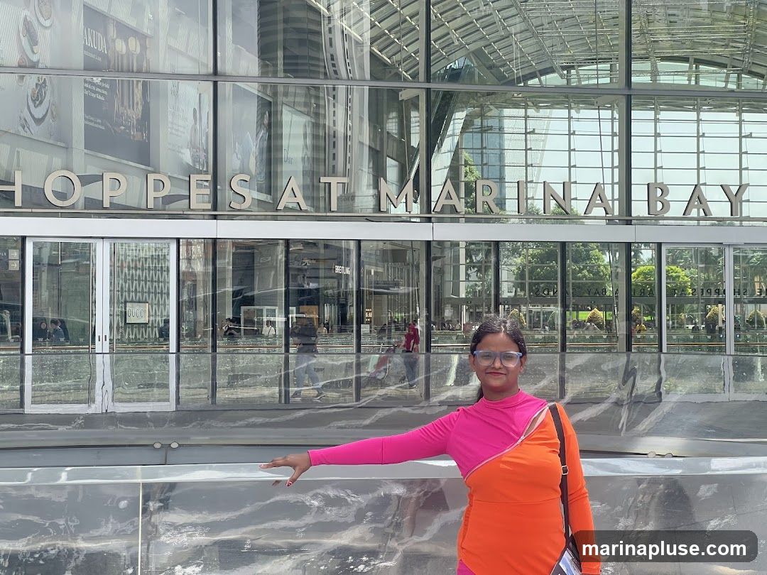 A woman stands in front of the entrance of The Shoppes at Marina Bay Sands. The building has a modern glass facade with large windows and the words "SHOPPES AT MARINA BAY" prominently displayed above the entrance. Reflections of trees and the sky are visible on the glass.