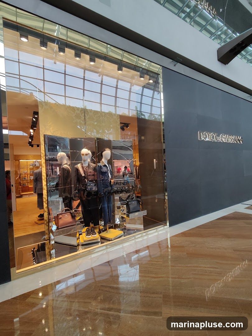 The exterior of a Dolce & Gabbana store with a large display window showcasing mannequins dressed in clothing and accessories. The store's name is visible in silver lettering on the dark facade to the right of the window. The interior of the store is visible through the window, with displays of merchandise. The floor in front of the store is highly reflective.