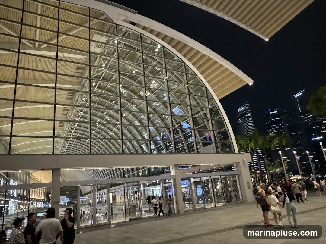 A wide shot of a modern building with a large glass facade and a curved roof, illuminated at night. Several people are visible walking in front of the building and entering through glass doors. In the background, city skyscrapers are lit up against the dark sky, with palm trees in the foreground.