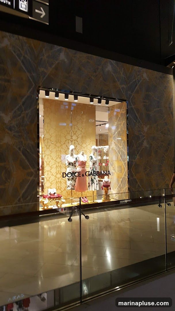 A well-lit storefront window display for Dolce & Gabbana, featuring mannequins dressed in the brand's clothing. The display is set against a backdrop of ornate gold wallpaper. The store's name is prominently displayed in large lettering on the glass.