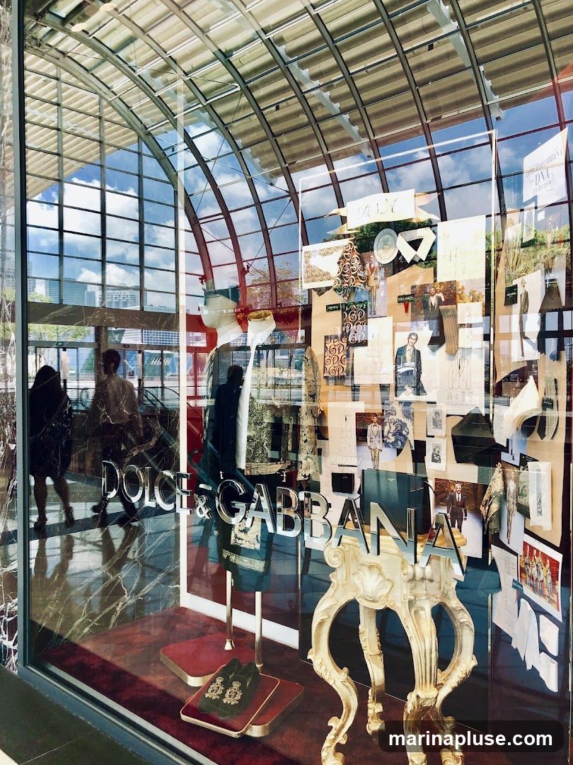 A reflection of a modern building with a curved glass ceiling is visible in the large display window of a Dolce & Gabbana store. The window showcases a collection of fashion sketches, fabric swatches, and clothing items, along with a pair of ornate loafers on display. The store's name, "DOLCE & GABBANA," is prominently displayed in large, metallic letters on the glass.