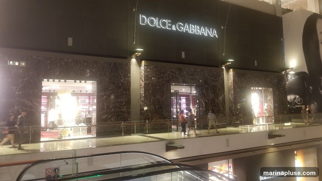 The exterior of a Dolce & Gabbana store is shown, featuring a dark marble facade with illuminated windows displaying merchandise. The brand name "DOLCE & GABBANA" is prominently displayed in large, glowing letters above the entrance. People are seen walking past the store on a walkway. A large advertisement featuring a woman's face is visible to the right of the store.
