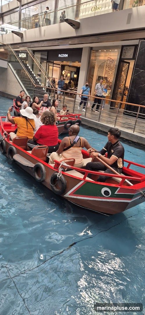 A group of people are enjoying a boat ride on a canal inside a modern shopping mall. The boat is red and brown with white accents. Several shops are visible in the background, including AVEDA and PAZZION. The mall has multiple levels with escalators and railings.
