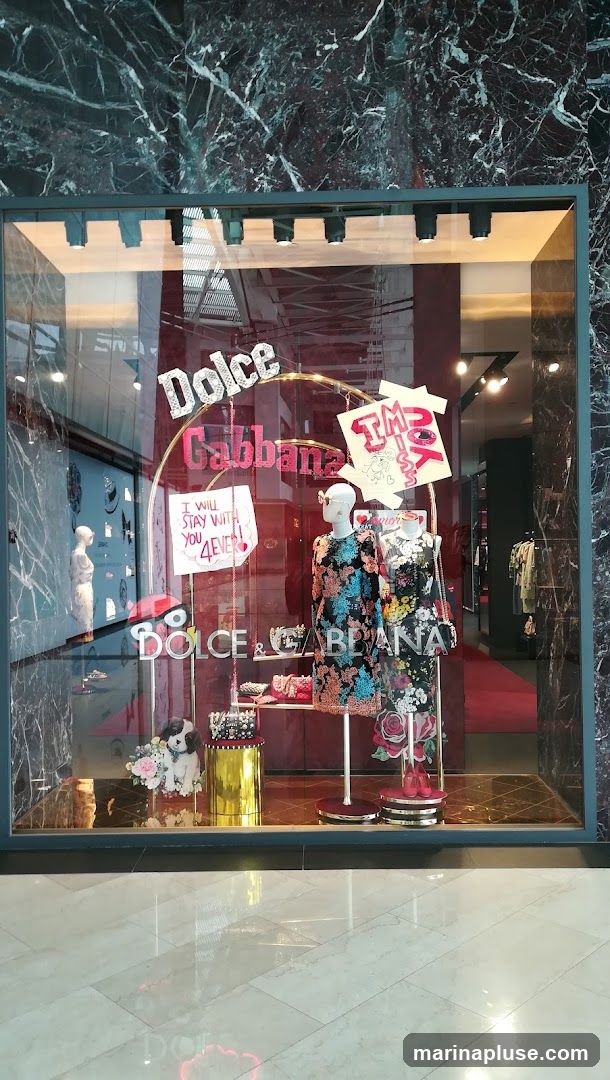 A store window display for Dolce & Gabbana featuring mannequins dressed in floral print dresses. The display includes the brand's logo, decorative elements like a dog with flowers, handbags, and signage.