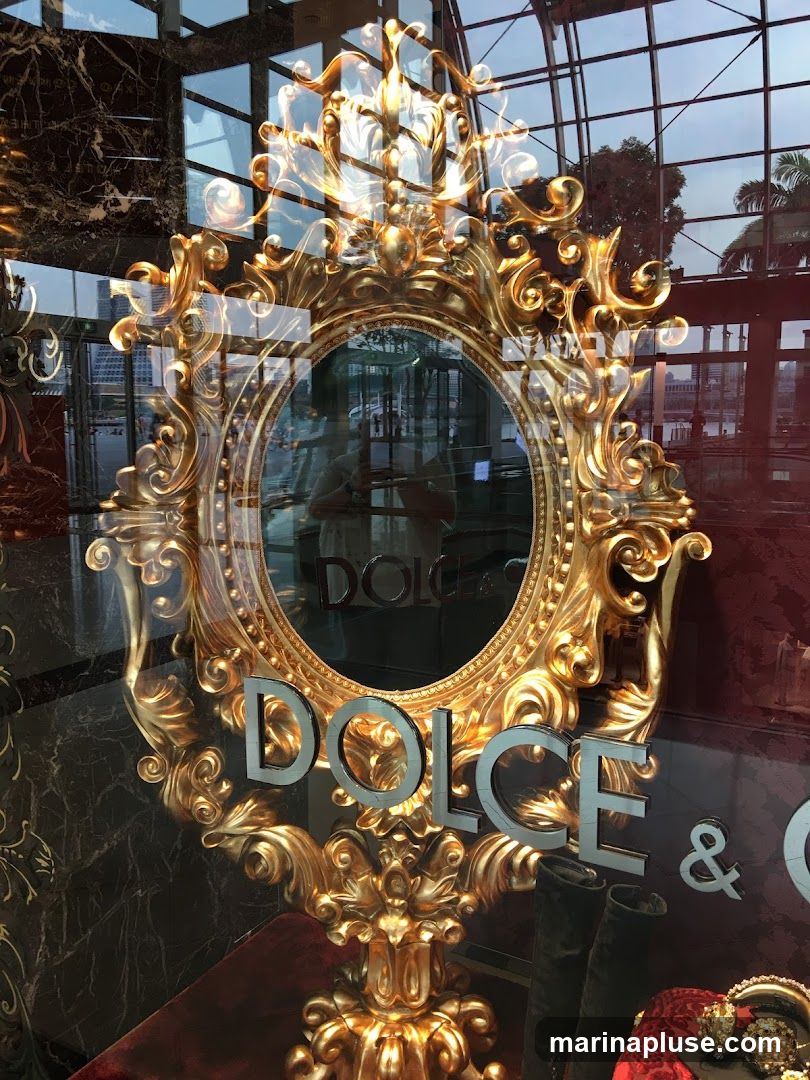 A ornate gold-framed mirror is displayed in a shop window. The words "DOLCE & GABBANA" are visible on the glass. Reflections of the interior and exterior are seen in the glass.