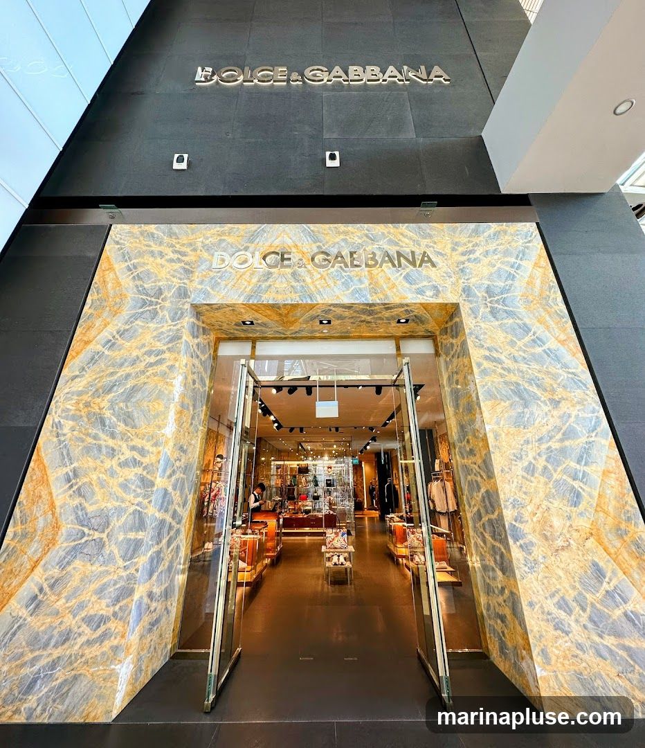 The exterior of a Dolce & Gabbana store with a grand entrance framed by marble. The store's name is prominently displayed above the entrance and on the facade. The doors are open, revealing a well-lit interior with merchandise displayed on shelves and racks.