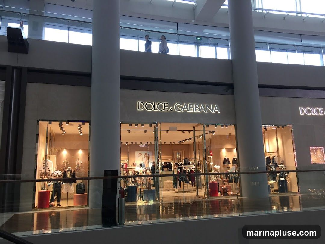 The exterior of a Dolce & Gabbana store in a modern shopping mall. The store features a large glass facade with illuminated signage above. Inside, mannequins display clothing and accessories. The reflection of the store and surrounding mall is visible on the polished floor.