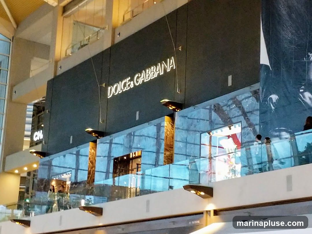 The image shows the exterior of a Dolce & Gabbana store located within a modern shopping mall. The store's name is prominently displayed in illuminated white lettering on a dark gray facade. Below the signage, a glass railing with reflective panels overlooks the mall's interior. The store appears to be on an upper level, with glimpses of merchandise and shoppers visible through the glass.