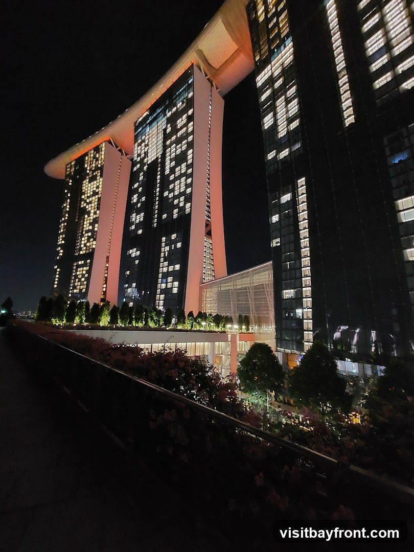 A low-angle night view of the Marina Bay Sands complex in Singapore. The iconic three towers, connected by the SkyPark, are illuminated with warm orange and white lights. The windows of the towers show many lit rooms, suggesting occupancy. In the foreground, a walkway is lined with lush greenery and flowering plants, also illuminated by subtle lighting. The overall scene conveys a sense of grandeur and urban vibrancy.