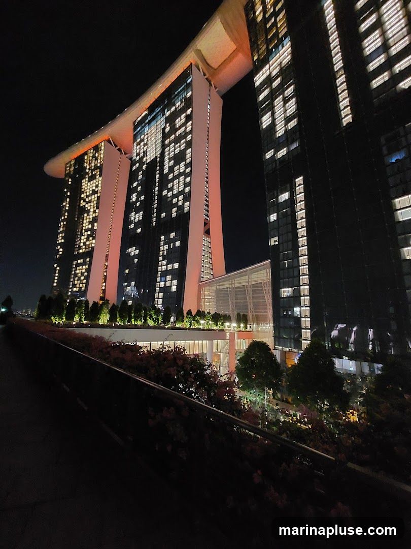 A low-angle night view of the Marina Bay Sands complex in Singapore. The iconic three towers, connected by the SkyPark, are illuminated with warm orange and white lights. The windows of the towers show many lit rooms, suggesting occupancy. In the foreground, a walkway is lined with lush greenery and flowering plants, also illuminated by subtle lighting. The overall scene conveys a sense of grandeur and urban vibrancy.