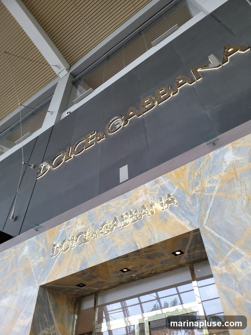 A close-up, low-angle shot of the exterior of a Dolce & Gabbana store. The brand's name is prominently displayed in large, gold, illuminated lettering on both a dark gray facade above and a marble-patterned archway below. The entrance appears to be through large glass doors, reflecting the interior and the sky outside.