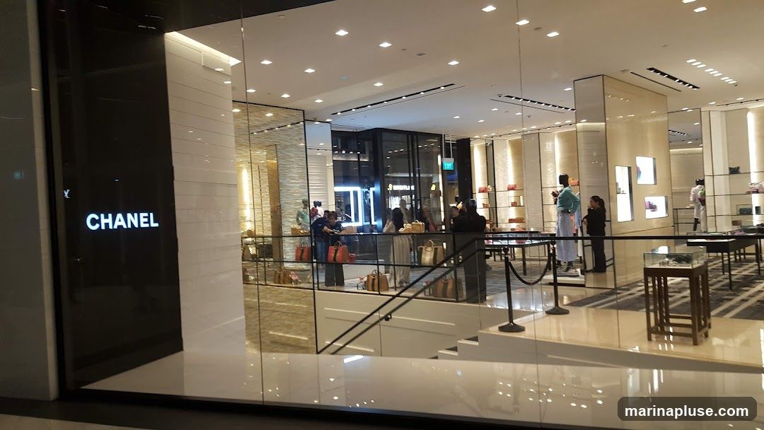 A view of the exterior of a Chanel store. The prominent "CHANEL" logo is visible on a dark facade to the left. The interior of the store is visible through large glass windows, showing displays of handbags, mannequins, and shelving units. Several customers are browsing inside.