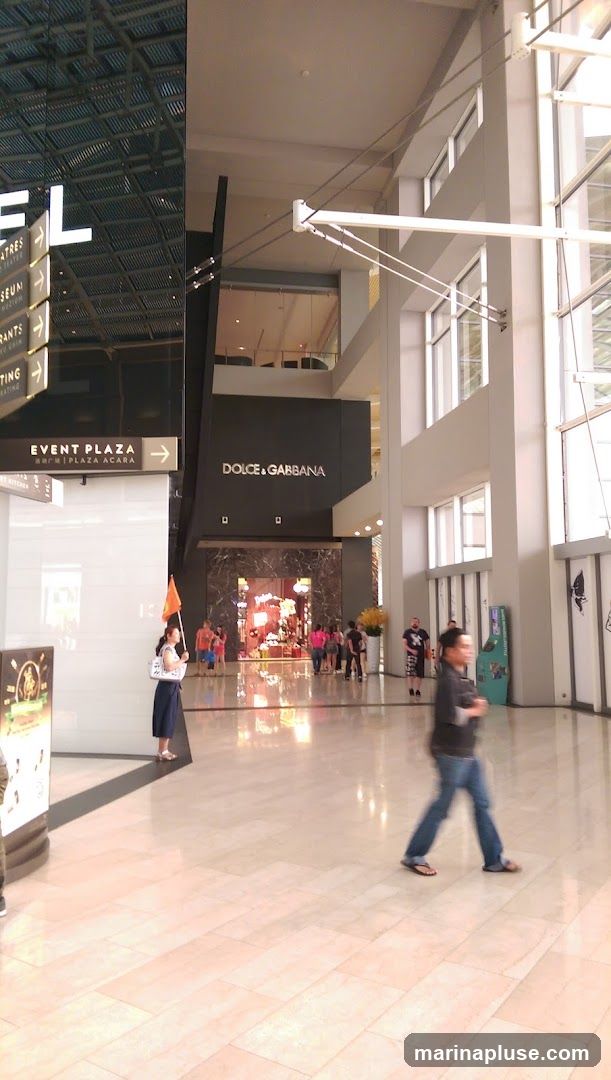 An interior view of a shopping mall with a Dolce & Gabbana store prominently displayed. The store has a dark facade with the brand name in white lettering above a marble-framed entrance. Inside, the store is brightly lit, showcasing merchandise. People are seen walking through the mall and in front of the store. Signs for "EVENT PLAZA ACARA" and other directions are visible on the left.