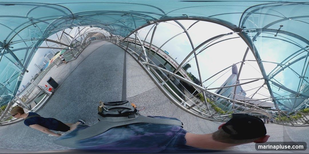 A 360-degree view of a modern, covered pedestrian bridge with a distinctive curved, glass-like roof structure. The bridge has a metal frame and a grey, speckled walkway. Several people are visible walking along the bridge, some in athletic wear. In the background, modern buildings and a body of water can be seen. The perspective is from someone walking on the bridge, with their back to the camera.