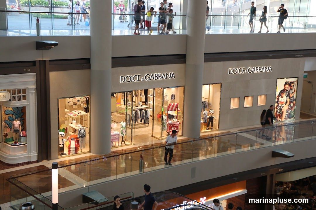 A view of the Dolce & Gabbana store in a mall. The store has a large sign with the brand name above its entrance. The windows display clothing and accessories. There are mannequins dressed in children's clothing. A man in a white shirt stands near the entrance.