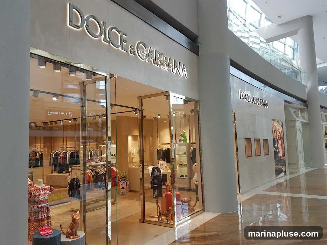 The exterior of a Dolce & Gabbana store with large glass windows and doors. The store's name is prominently displayed in illuminated letters above the entrance. Inside, clothing racks and displays of merchandise are visible.