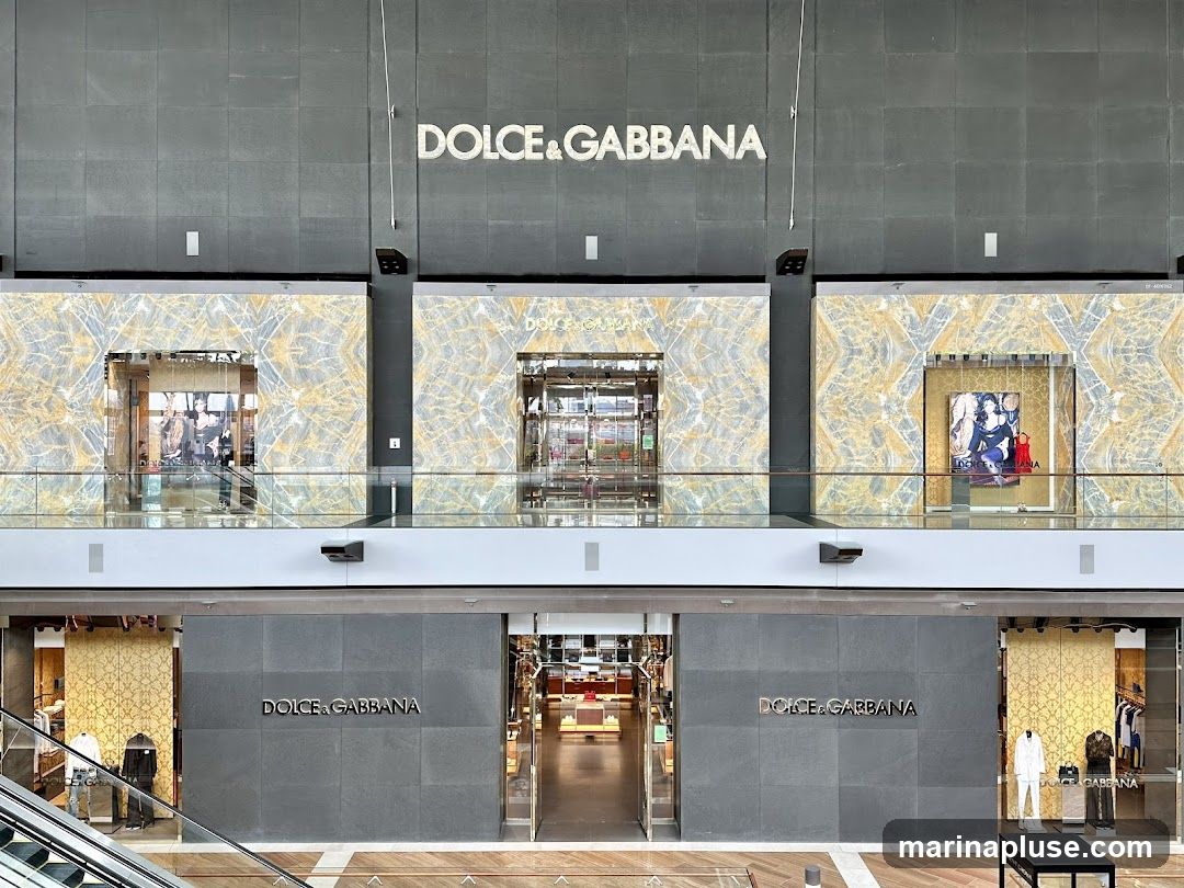 The exterior of a Dolce & Gabbana store with large display windows and the brand name prominently displayed above the entrance. The facade features a modern design with marble-like accents and sleek signage. Inside, clothing and accessories are visible on display racks and mannequins.