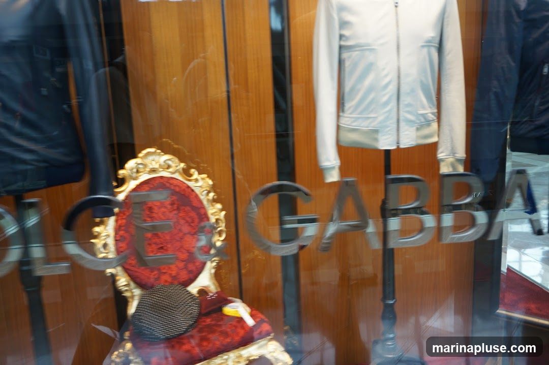 A reflection on a shop window shows a white jacket on a mannequin, a dark jacket on another mannequin, and a decorative red chair with gold trim. The words "DOLCE & GABBANA" are visible as metallic lettering on the glass.
