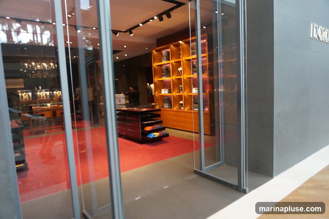 A view through a glass storefront into a well-lit retail space. The interior features red carpeting, wooden display shelves filled with merchandise, and a central display table. A sign is visible on the exterior gray wall to the right.