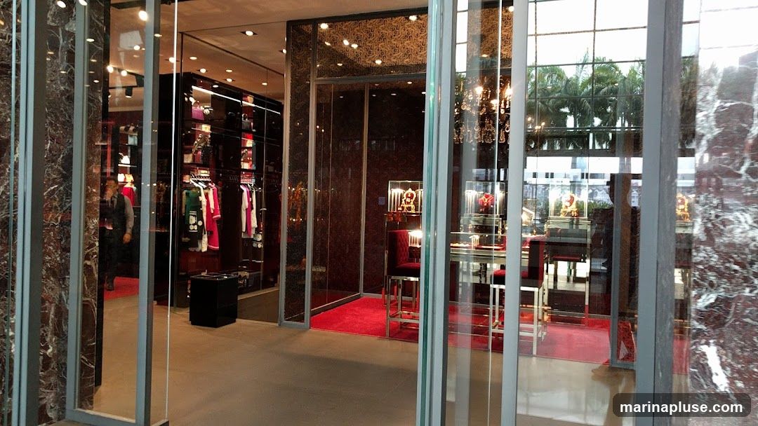 A view into a high-end retail store, showcasing clothing and jewelry displays. The interior features dark, opulent decor with red accents, including plush seating and red carpets. Racks of designer clothing and illuminated display cases filled with jewelry are visible. The store appears to have large glass windows and doors, reflecting the exterior environment.