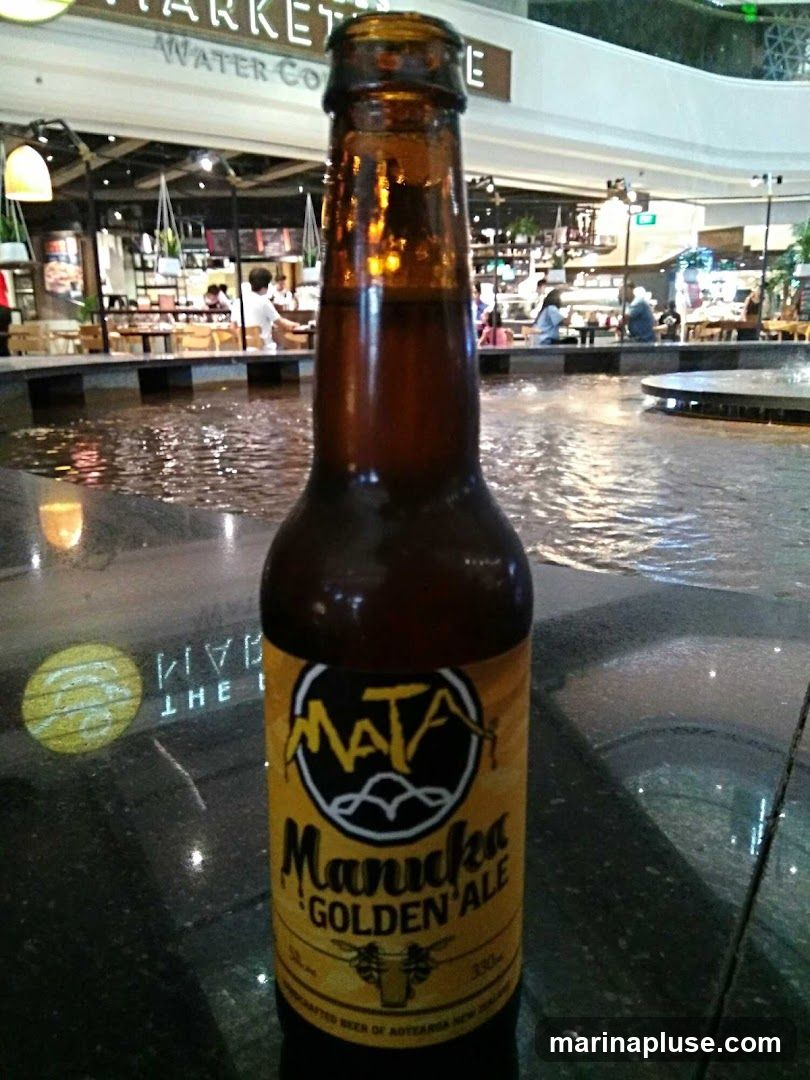 A close-up of a brown glass bottle of Manuka Golden Ale, with the label clearly visible. The bottle is placed on a dark, reflective surface, possibly a table, in front of a blurred background of a marketplace or food court with a water feature. The label features the brand name "MATA" and "Manuka GOLDEN ALE" in yellow and black text, along with an illustration of a cow and bees. The background shows people dining and shopping in a bright, indoor environment.