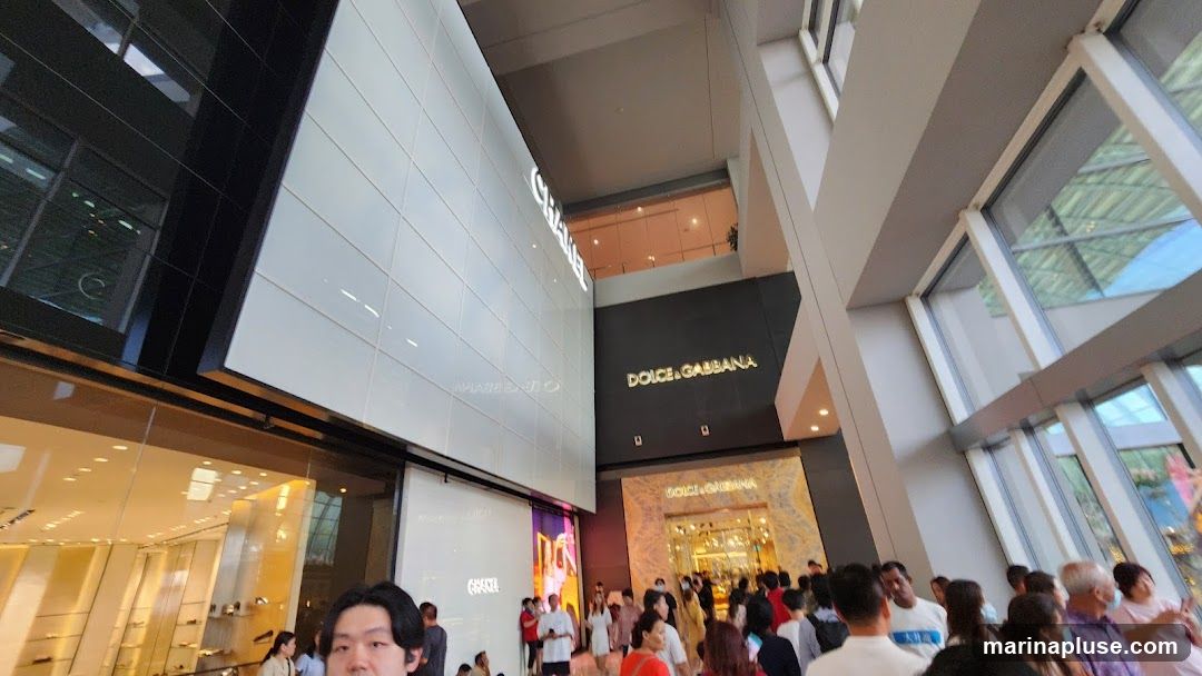 A wide shot of the interior of a modern shopping mall, with a focus on the storefronts of Chanel and Dolce & Gabbana. The Chanel store features a large, white tiled facade with the brand name illuminated in large, white letters. Below it, a glass storefront displays merchandise. The Dolce & Gabbana store is visible to the right, with its name in gold lettering on a dark background, and an ornate entrance below. A crowd of shoppers is gathered in front of the Dolce & Gabbana store. The mall's architecture includes large windows and a high ceiling, with natural light filtering in.