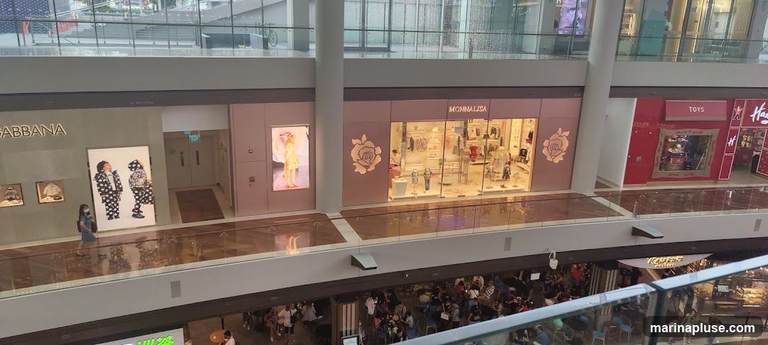 An elevated view of several storefronts in a modern shopping mall. On the left, a store with 'DOLCE & GABBANA' signage displays artwork and clothing. Next to it is a store with a large digital display featuring a child in a dress. Further to the right is a pink storefront with the name 'MONNALISA' and rose motifs, showcasing children's clothing. To the far right, a red storefront labeled 'TOYS' is visible, with a whimsical mirror and signage for 'Harrods'. Below these, a busy food court area is visible with many patrons seated at tables.