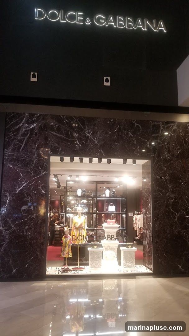The exterior of a Dolce & Gabbana store. The store has a dark marble facade and a large display window. Inside the window, mannequins are dressed in colorful clothing, and handbags are displayed on pedestals. The store's name, "DOLCE & GABBANA," is illuminated in large letters above the window.
