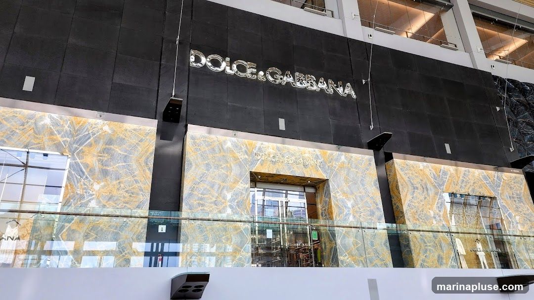 The exterior of a Dolce & Gabbana store. The facade features a large black stone wall with the store's name in prominent silver lettering. Below this, the entrance is framed by marble with gold veining. Glass doors lead into the store, where merchandise is visible. A glass railing is in the foreground.
