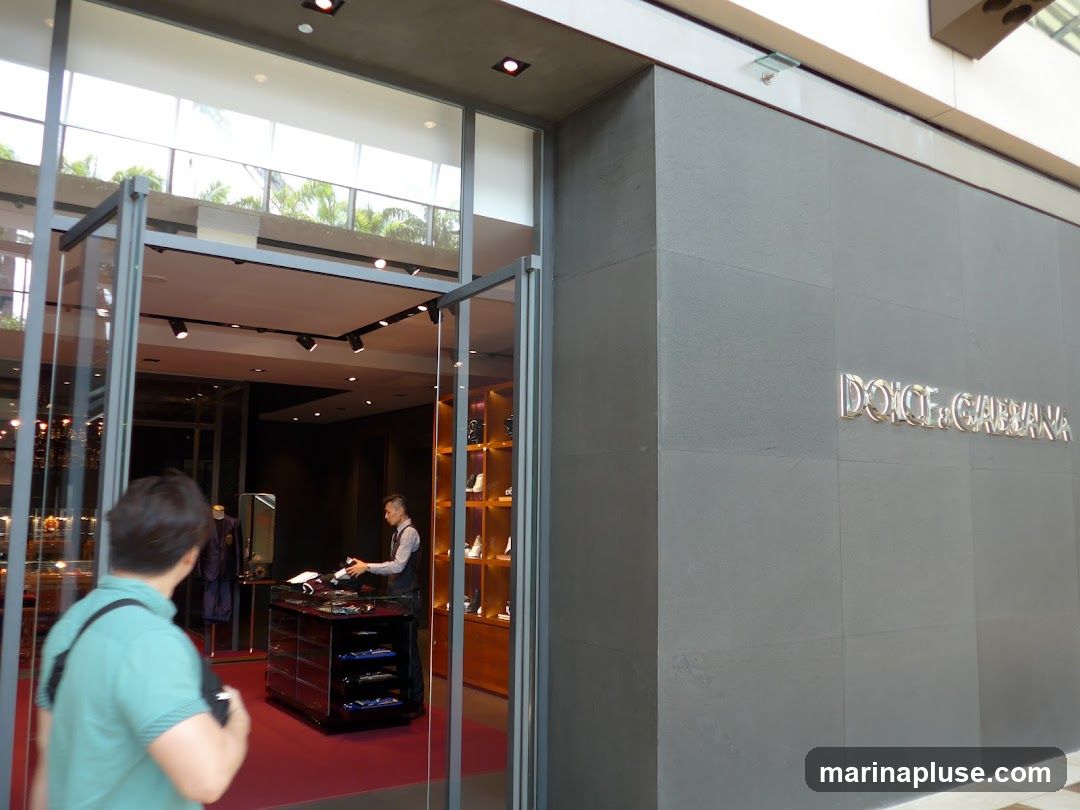 The exterior of a Dolce & Gabbana store. The store has a large gray facade with the brand name "DOLCE & GABBANA" in silver lettering. To the left, there are glass doors and windows revealing the interior of the store, where a staff member is attending to a customer. The interior displays clothing and accessories on shelves and racks.