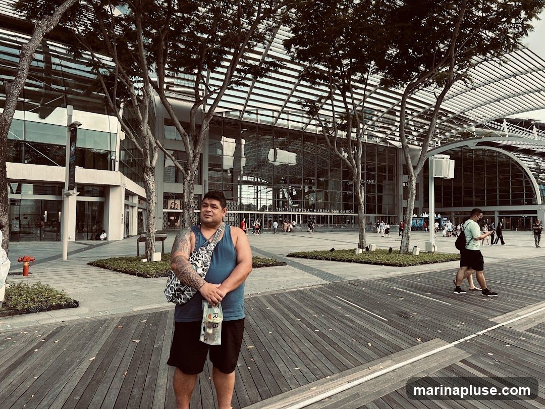 A man stands on a wooden boardwalk, looking to his left. Behind him is a large, modern building with a glass facade, identified by signage as 'THE SHOPPES AT MARINA BAY SANDS'. Several trees are planted in front of the building. Other people are visible walking in the distance.