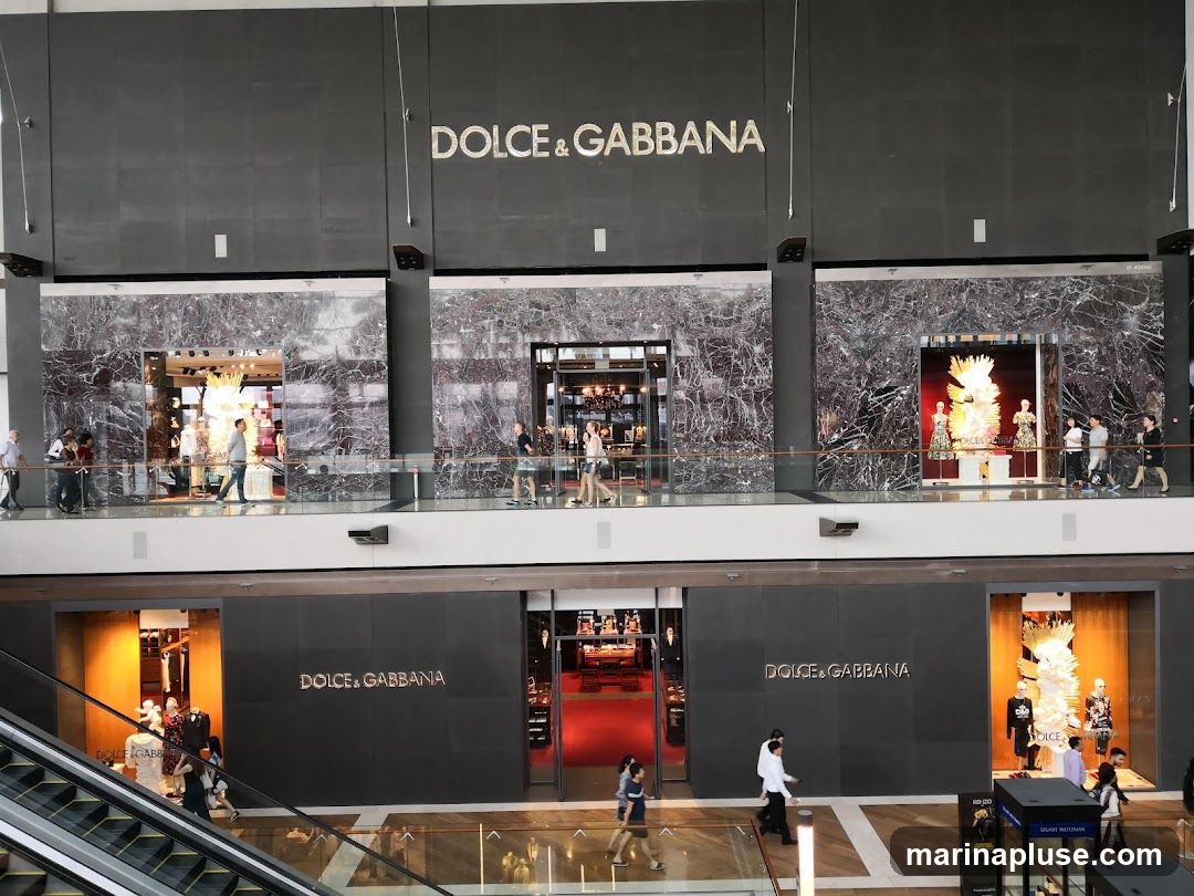 A wide shot of the Dolce & Gabbana store on two levels of a mall. The upper level has three large display windows with marble accents and mannequins dressed in elaborate outfits. The Dolce & Gabbana logo is prominently displayed above these windows. The lower level features two entrances, each with the Dolce & Gabbana logo above. One entrance leads to a store interior with red carpeting, while the other has a display window with mannequins. An escalator is visible on the left side of the lower level.