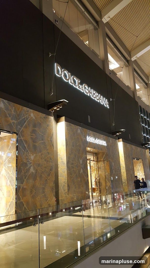The exterior of a Dolce & Gabbana store with illuminated signage. The facade features marble accents and large glass entrances. The store is located within a modern indoor shopping mall, with a reflective floor and a glass railing in the foreground.