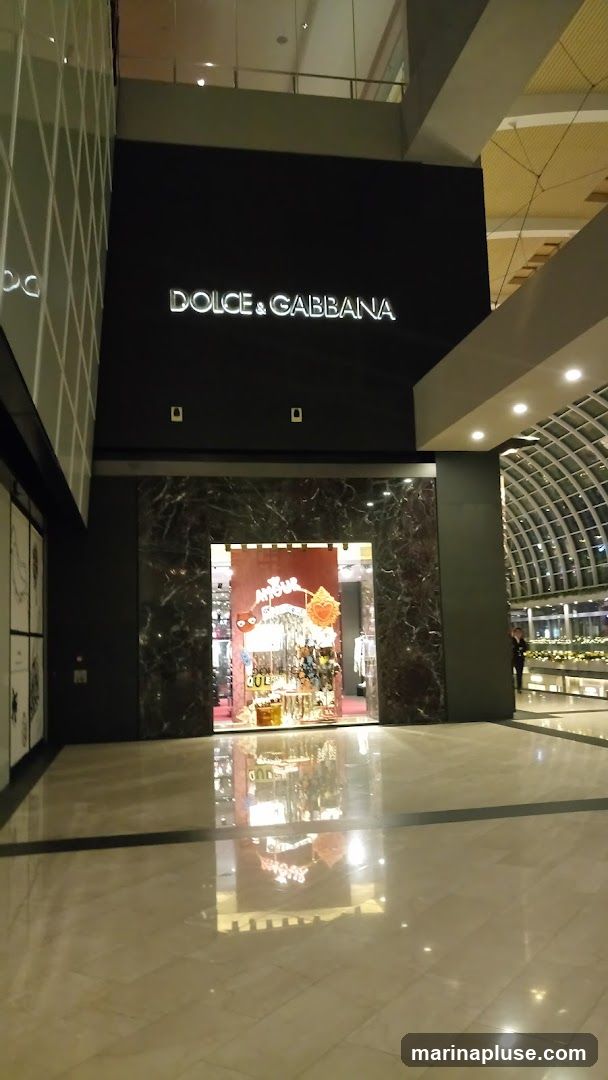 The exterior of a Dolce & Gabbana store is shown, with the brand name illuminated above the entrance. The storefront features a large display window showcasing merchandise. The interior of the store is visible through the window, with displays of various items. The floor in front of the store is highly reflective, mirroring the store's facade and lighting.
