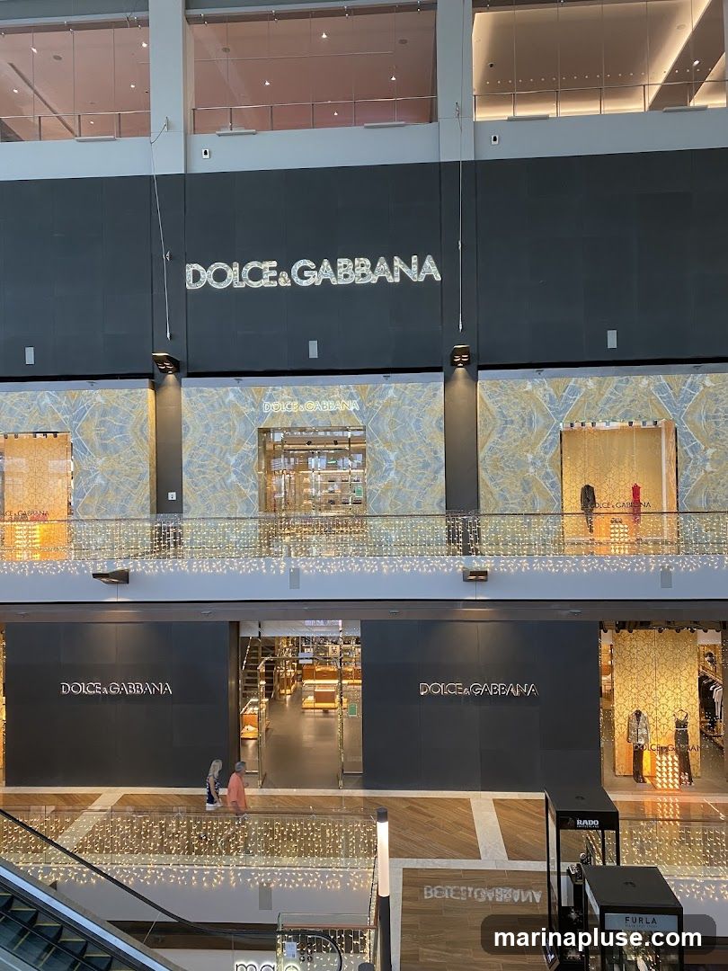 A multi-level view of a Dolce & Gabbana store within a shopping mall. The store features prominent signage with the brand name on the facade. The interior is visible through large glass entrances, showcasing display cases and merchandise. Decorative lights are strung across the balconies.