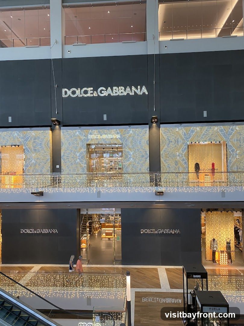 A multi-level view of a Dolce & Gabbana store within a shopping mall. The store features prominent signage with the brand name on the facade. The interior is visible through large glass entrances, showcasing display cases and merchandise. Decorative lights are strung across the balconies.