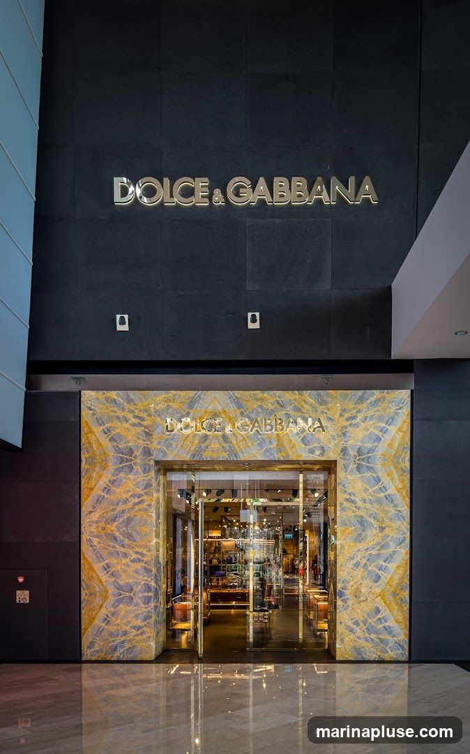 The exterior of a Dolce & Gabbana store. The entrance is framed by marble with gold veining. The store name is displayed in gold lettering above the entrance and on the dark facade above. Through the glass doors, merchandise and displays are visible inside.