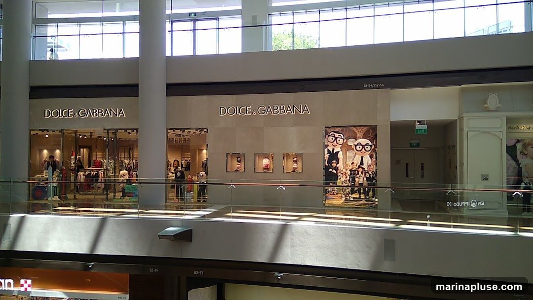 The image shows the exterior of a Dolce & Gabbana store located in a mall. The storefront features the brand's name prominently displayed above the entrance. Inside, clothing and accessories are visible on display racks and mannequins. To the right of the main entrance, there is a large advertisement featuring cartoon characters and children. The store is situated on an upper level of the mall, with a glass railing in the foreground.