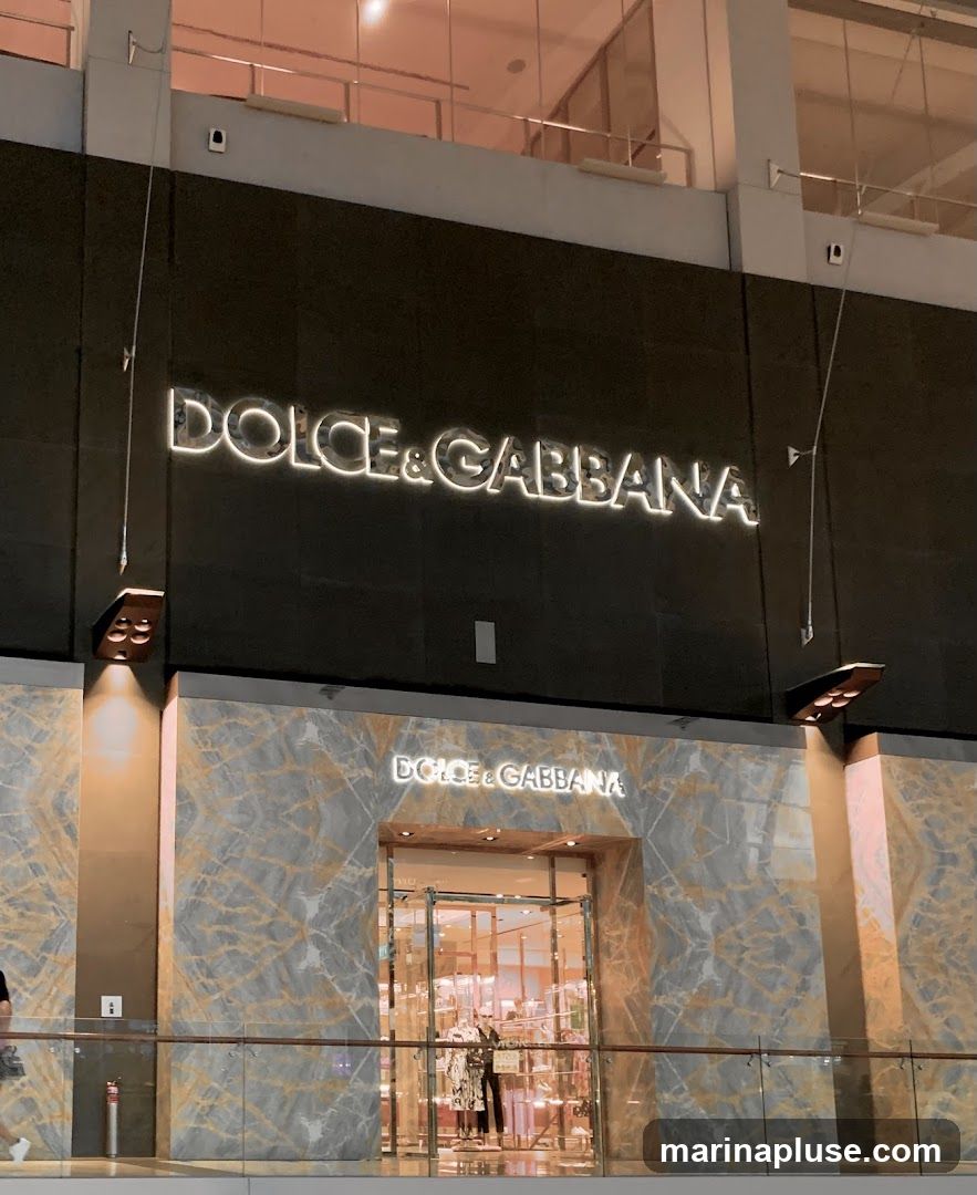 The exterior of a Dolce & Gabbana store. The store has a marble facade with the brand name "DOLCE & GABBANA" displayed in large illuminated letters above the entrance. The entrance is a glass door with a metal frame, and inside, mannequins displaying clothing can be seen.