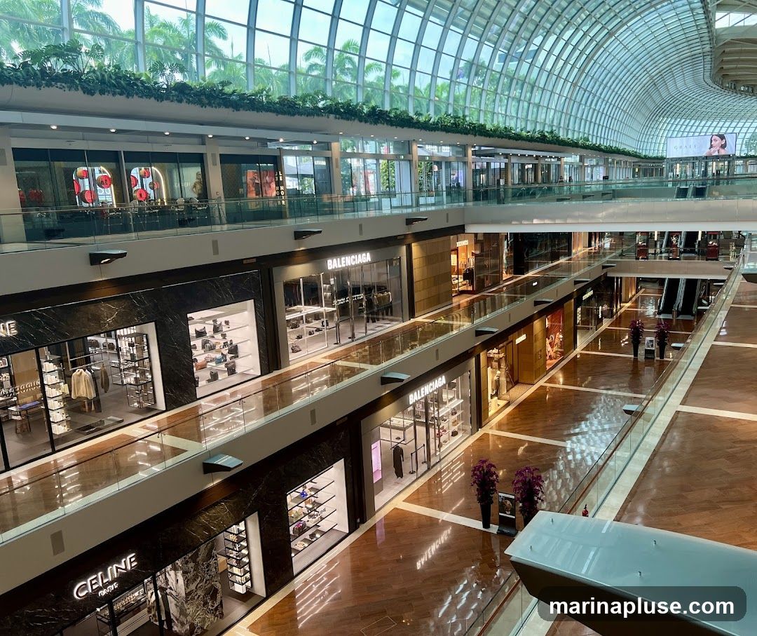 An interior view of a multi-level shopping mall with a glass ceiling and lush greenery. Several high-end stores are visible, including Celine and Balenciaga, displaying their merchandise. Escalators and walkways connect the different levels, and the polished wooden floors reflect the ambient light.